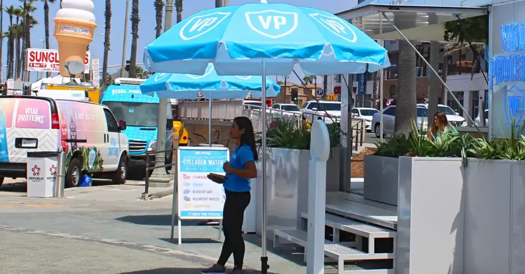 A woman stands under a large blue umbrella with VP logos, next to a Vital Proteins booth on a sunny street with palm trees, parked vans, and a large ice cream cone sculpture in the background.