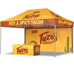 A yellow and brown taco stand setup with a canopy labeled HOT & SPICY TACOS. The booth and backdrop feature taco graphics, text, and a cactus illustration, creating a festive, inviting atmosphere.