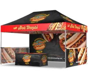 A pop-up canopy tent decorated with images of hot dogs and the text Hot Dogs! The tent, table, and counter feature matching branding with colorful hot dog graphics.