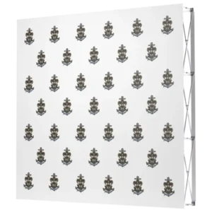 A white step and repeat banner featuring a repeating pattern of a navy and gold anchor emblem with a shield and ribbon design. The emblems are evenly spaced across the banner.