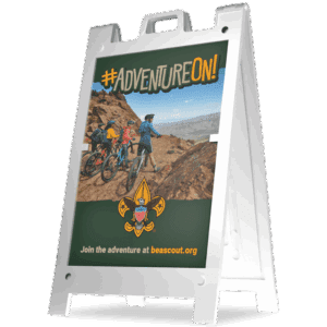 A sidewalk sign displays a photo of four kids on mountain bikes riding a rocky trail. The sign reads #AdventureOn! and Join the adventure at beascout.org with the Boy Scouts of America logo.