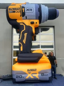 A person stands next to a giant inflatable DeWalt drill prop, which is black and yellow with 20V MAX XR 8Ah and BRUSHLESS labels, in front of a gray building with large windows.
