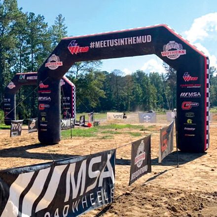 An outdoor off-road event entrance with large black inflatable arches displaying “#MEETUSINTHEMUD” and various sponsor logos, surrounded by trees and fenced pathways on a sunny day.