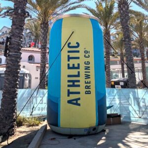 A large inflatable can with the label Athletic Brewing Co. stands outdoors surrounded by palm trees and buildings under a sunny sky.