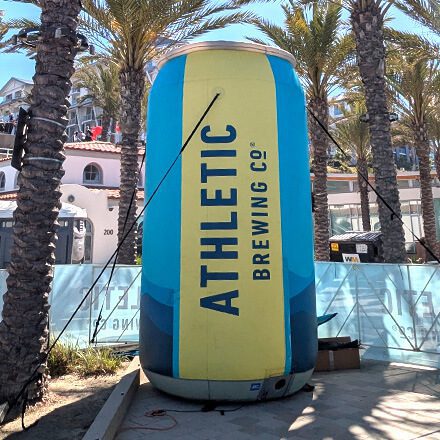 A large inflatable can with the label Athletic Brewing Co. stands outdoors surrounded by palm trees and buildings under a sunny sky.