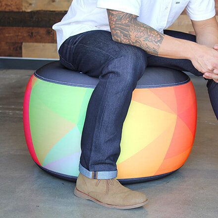 A person with tattooed arms, wearing a white shirt, dark jeans, and tan shoes, sits on a round, colorful ottoman with a geometric rainbow pattern in a modern room with a concrete floor.