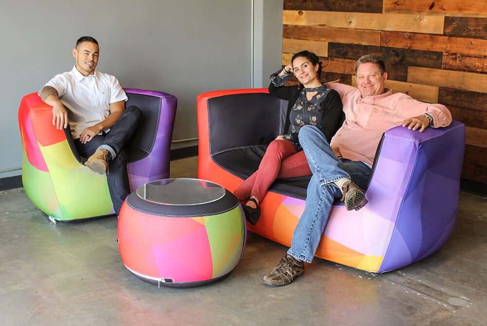 Three people sit and relax on colorful, modern chairs with geometric patterns. A man sits sideways in an armchair, while a woman and another man sit together on a matching sofa. A round table is in front of them.