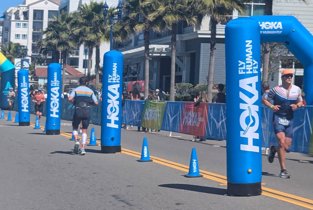 Two athletes run on a sunny street during a race, passing blue HOKA-branded inflatable arches. Spectators watch behind barriers, and palm trees line the road near modern buildings.