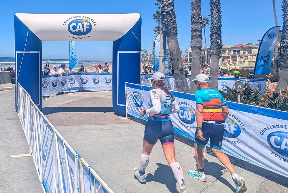 Two athletes in athletic gear approach a finish line arch at an outdoor event by the beach, with palm trees and spectators in the background on a sunny day.