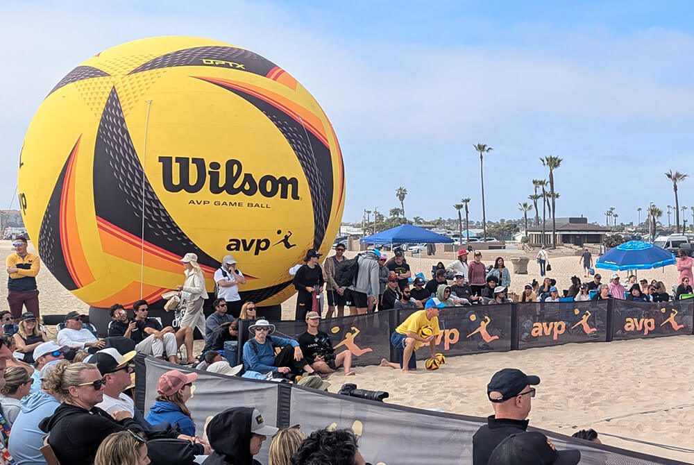 A beach volleyball event with a large, inflatable Wilson volleyball displayed behind a crowd of spectators. People are sitting and standing on the sand, and palm trees are visible in the background under a partly cloudy sky.
