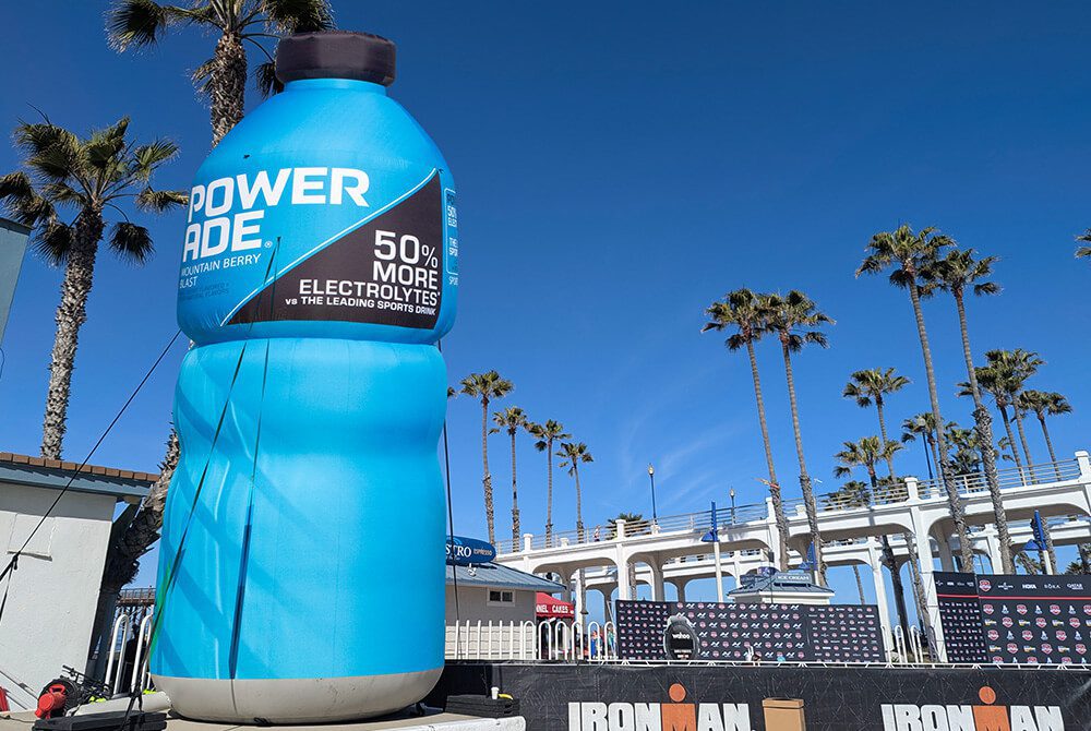 A giant inflatable blue Powerade bottle stands outdoors near palm trees and a stage with Ironman branding, under a clear blue sky.