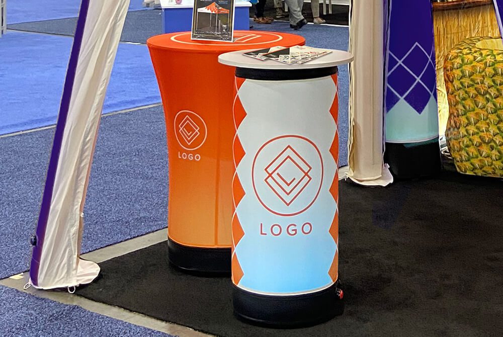 Two cylindrical display stands with geometric “LOGO” designs, one orange and one blue, are set up on a black carpet at an event. A small stack of flyers and a brochure holder are placed on top.