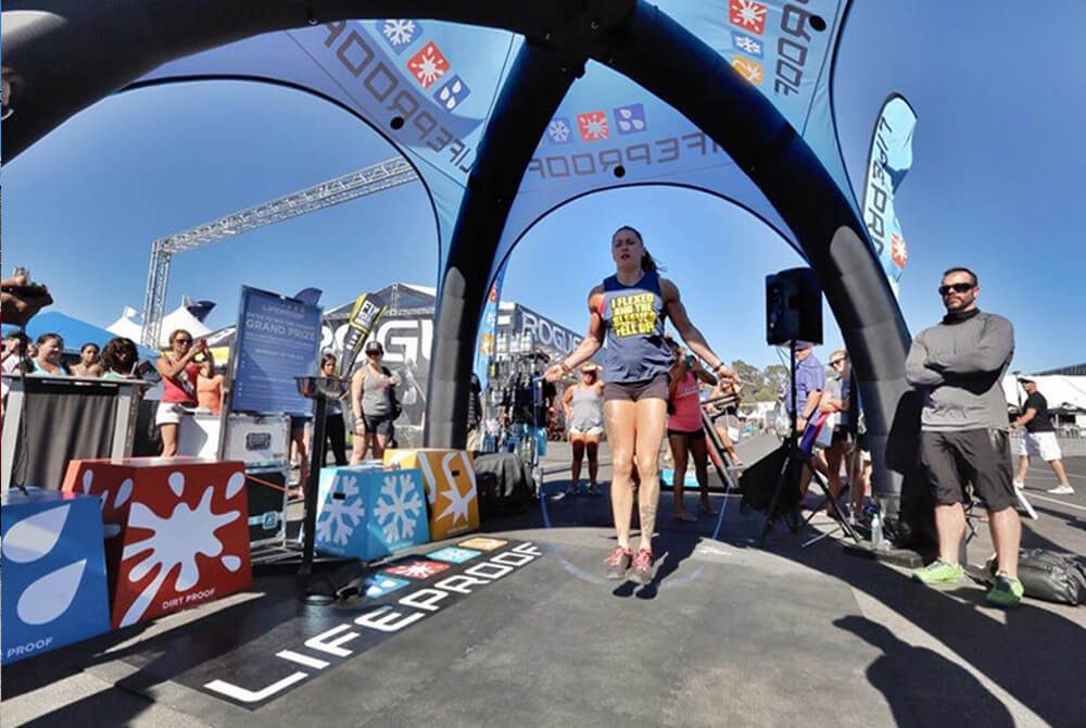 A woman in athletic wear jumps rope under a branded inflatable arch at an outdoor event, while spectators watch and the sun shines in a clear blue sky.