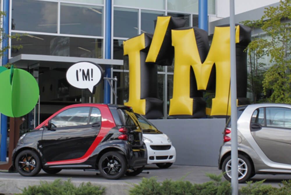 Two small cars parked outside a modern glass building, one red and black, the other silver. Large yellow and black inflatable letters spell “I’M,” with a speech bubble near the cars also saying “I’M!”.