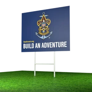 A blue sign with the Sea Scouts logo, website SeaScout.org, and the slogan Build an Adventure stands on a white frame on green grass.