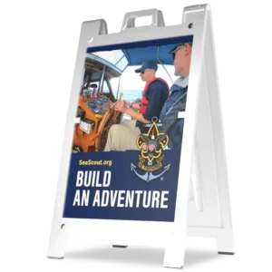 A white sandwich board sign displays two people steering a boat, with the Sea Scouts logo and the words SeaScout.org BUILD AN ADVENTURE in bold letters.