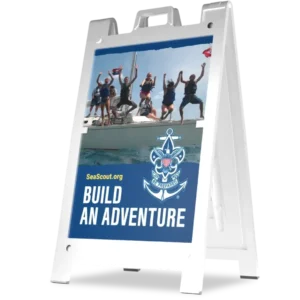 A white sandwich board sign displays a photo of six excited young people on a sailboat, with the Sea Scouts logo and the text “SeaScout.org BUILD AN ADVENTURE” underneath the image.