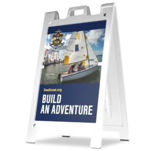 A sandwich board sign with an image of youths sailing a small boat, the Sea Scouts logo, and text reading SeaScout.org BUILD AN ADVENTURE against a blue background. More sailboats are visible in the background.