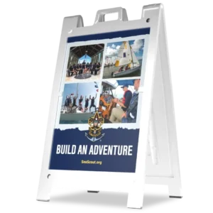 A white A-frame sign displays four photos of youth boating activities above the Sea Scouts logo and the text Build an Adventure with SeaScout.org on a blue background.