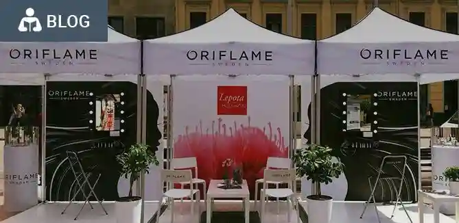Outdoor Oriflame event booth with three white tents, branded banners, chairs, tables, and potted plants, set up in a sunny area. A BLOG label appears in the top left corner.