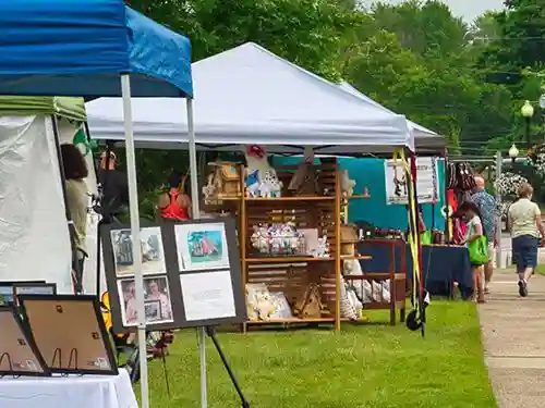 Outdoor art and craft fair with people walking by booths displaying framed photos, pillows, and handmade items under white and blue tents on a grassy area. Trees are visible in the background.