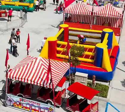 A colorful outdoor carnival features red-and-white striped tents, an inflatable obstacle course with children playing, and a small train ride surrounded by people walking and enjoying the event.