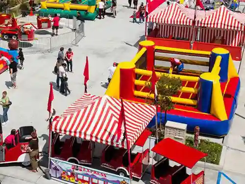 Aerial view of a lively outdoor fair with red-and-white striped tents, inflatable bounce houses, people walking, and a small train ride, all set up on a sunny day.