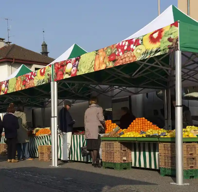 People shopping at an outdoor market stand with green and white striped tents and displays of oranges, lemons, and other produce stacked on crates under a sunny sky.