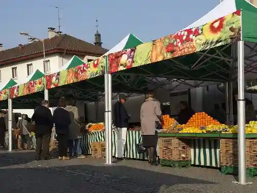 People shop for fresh fruits and vegetables at outdoor market stalls covered with green and white tents in a European town. The stalls have produce stacked on tables and crates underneath.