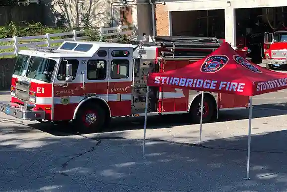 A red and white fire truck labeled E1 is parked outside a fire station. In front of it is a red canopy tent with Sturbridge Fire written on it. Another fire truck is visible inside the station.