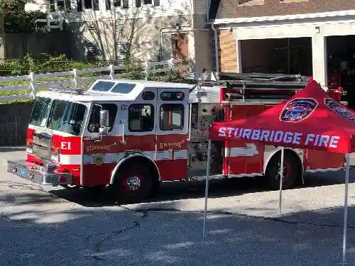 A red and white fire truck labeled E1 is parked next to a red tent that reads STURBRIDGE FIRE outside a building on a sunny day.