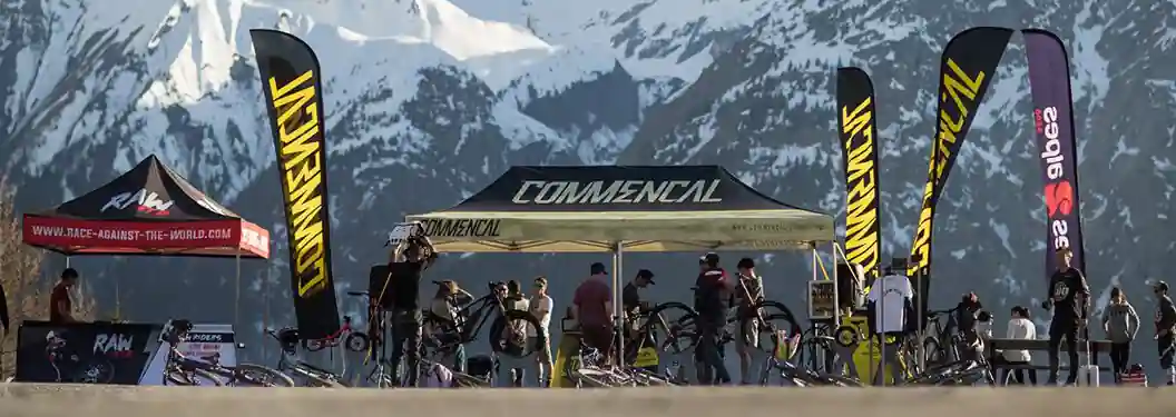 A group of people gather under branded tents with mountain bikes, set against snow-capped mountains. Flags with COMMENCAL text are visible, suggesting a biking event or expo in an outdoor alpine setting.