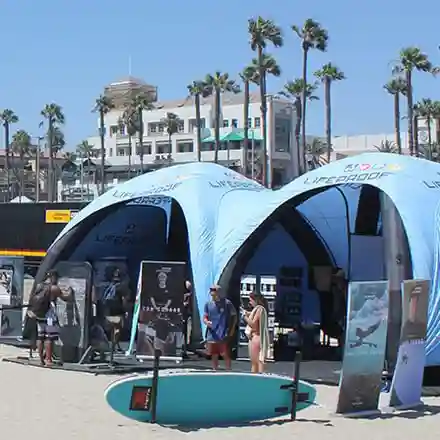 Outdoor event on a sunny beach with palm trees, featuring two large blue canopy tents, display stands, and several people walking or browsing. A surfboard is propped up in the foreground. Buildings are visible in the background.