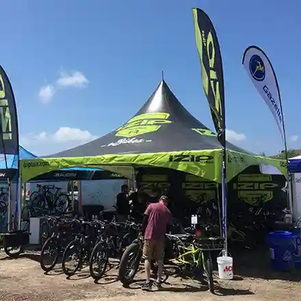 People stand under a large black and green tent displaying bikes at an outdoor event. Several bicycles are lined up in front of the tent, and branded flags are visible under a sunny sky.