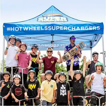 A group of kids and adults wearing helmets pose with skateboards and scooters under a blue tent labeled #HotWheelsSuperChargers during an outdoor event. Everyone looks excited and happy.