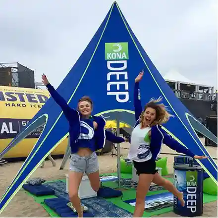Two women are jumping and smiling in front of a blue Kona Deep-branded tent at a beach event, with promotional items and other branded setups visible around them.