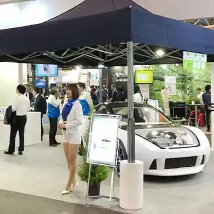 A woman in a white outfit stands under a canopy next to a white car with its hood open at an indoor exhibition, with several people and display booths in the background.