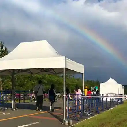 People walk under white tents set up along a fenced pathway outdoors, with a rainbow arching across a cloudy sky in the background.