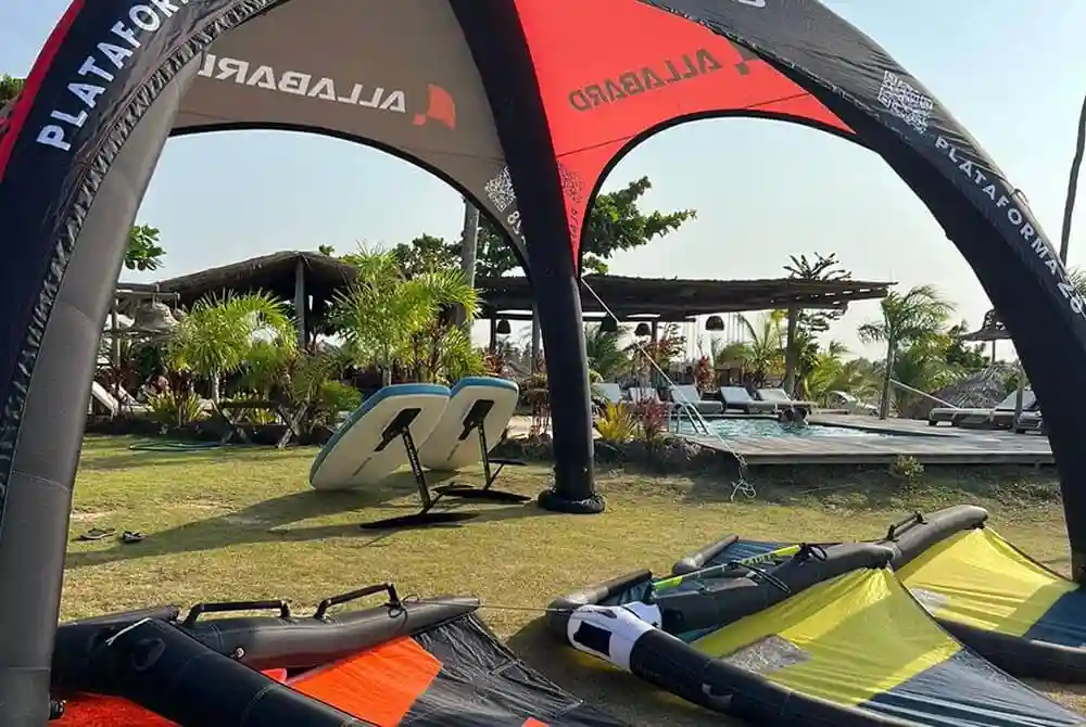 Large inflatable ALLABRAND tent on grass near a pool with palm trees. Two e-foils and surf equipment are set up outside, with people relaxing in the background under shaded seating areas.