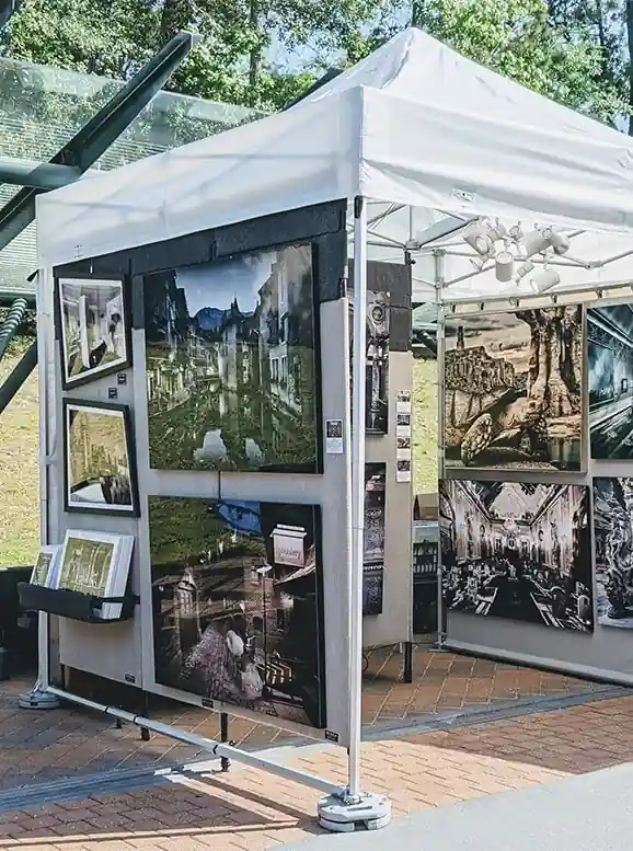 Outdoor art booth displaying framed cityscape and architectural photographs, set up under a white canopy tent on a brick walkway with greenery in the background.