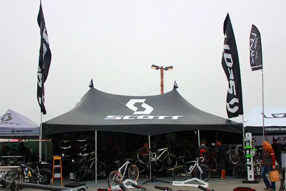A large black tent with the SCOTT logo and name, two tall SCOTT flags, and several bicycles displayed underneath. People are walking around, viewing the bikes at an outdoor event.
