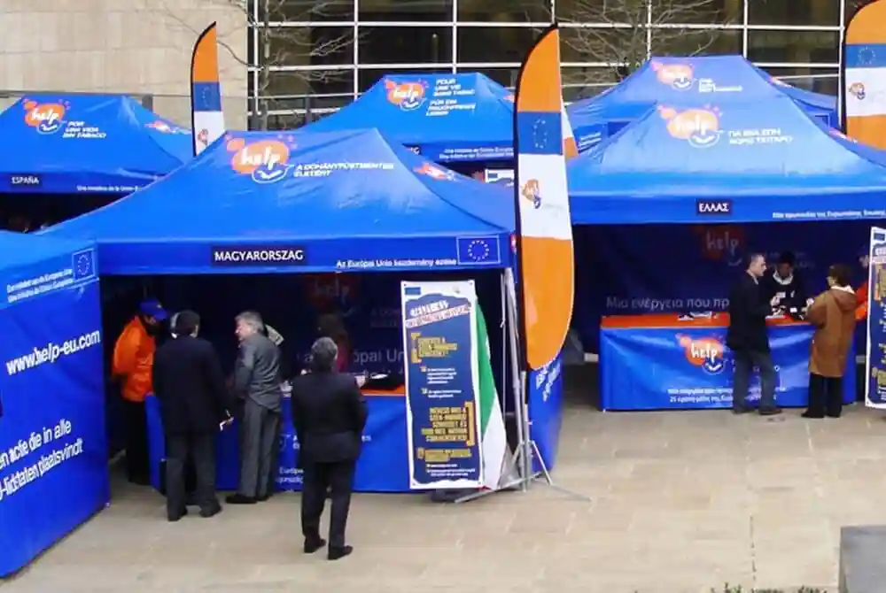Several blue information tents with European Union flags and signs are set up outdoors. People are standing near or inside the tents, talking and reading information boards. The setting appears to be an event or fair.