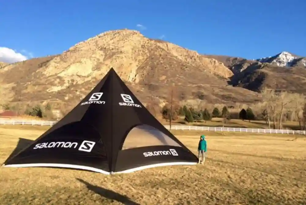 A person stands next to a large black and white Salomon tent on an open grassy field, with mountains and a blue sky in the background.