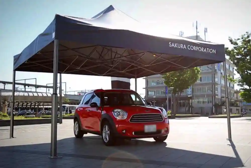 A red car is parked under a large black canopy tent labeled SAKURA CORPORATION in an open, sunlit urban area with buildings and trees in the background.