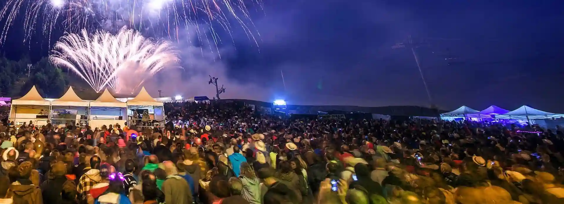 A large crowd gathered at an outdoor festival at night watches a bright fireworks display in the sky, with tents and stage lights visible in the background.