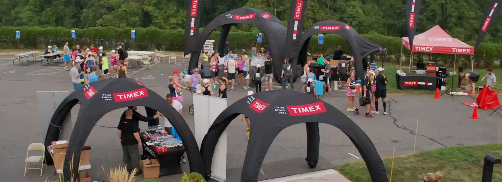 Outdoor event with people gathered under black inflatable tents branded with TIMEX. Tables, merchandise, and event materials are set up around the area. Green trees and parked cars are visible in the background.