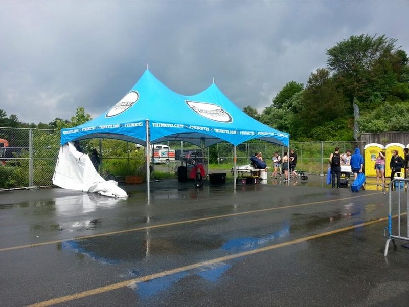 A 20x40 Printed Marquee Tent stands on wet pavement under a cloudy sky, its reflection visible in puddles. Some people gather nearby, yellow portable toilets are in the background, and part of the tent appears collapsed.