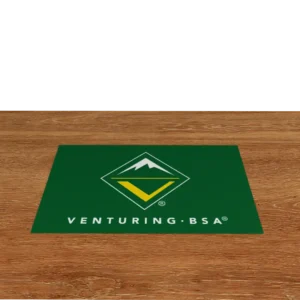 A green mat with the Venturing BSA logo, featuring a mountain inside a yellow and green diamond shape, lies on a wooden floor. VENTURING BSA is written in white below the logo.