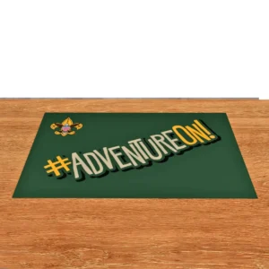 A green mat on a wooden surface features the Boy Scouts of America logo and the phrase #ADVENTUREON! in bold, stylized letters with yellow and white accents.