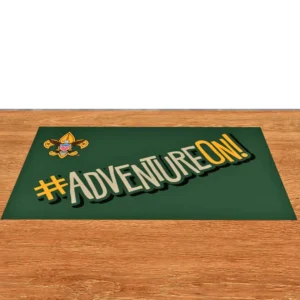 A green mat on a wooden floor displays the Boy Scouts of America emblem and the phrase #ADVENTUREON! in bold, stylized letters with yellow and white accents.
